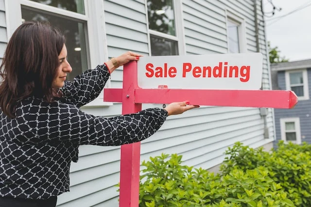 a woman hanging a 'sale pending' sign post in front of a home